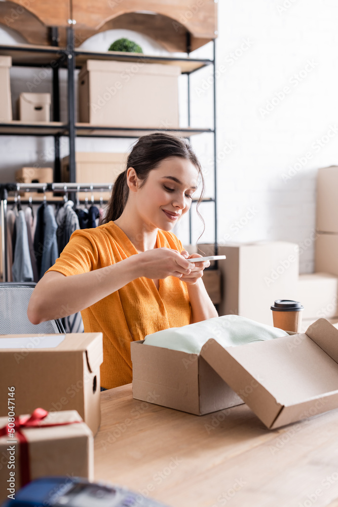 Smiling proprietor taking photo on clothes in carton box in online web store. Smiling proprietor taking photo on clothes in carton box in online web store.