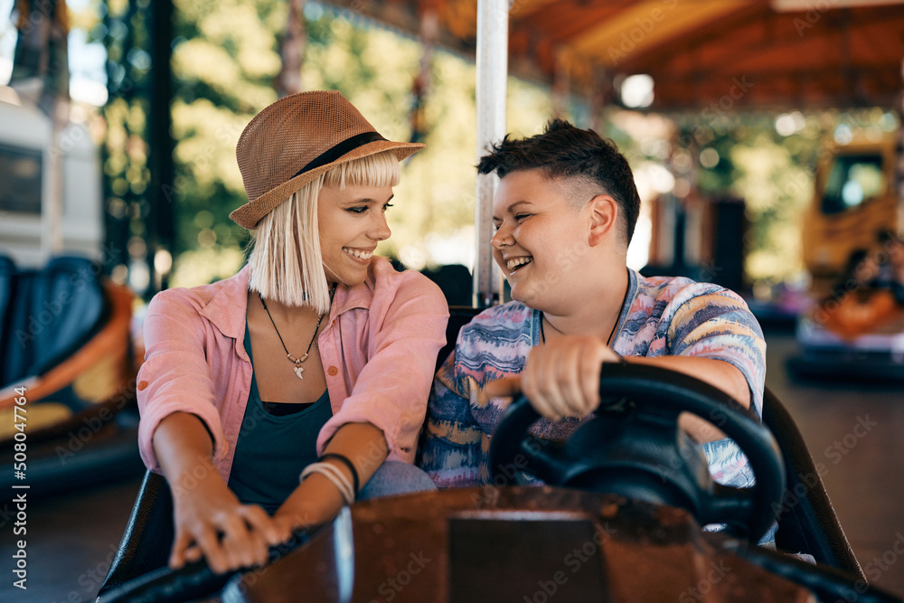 Happy lesbian couple talks while driving bumper car at amusement park.