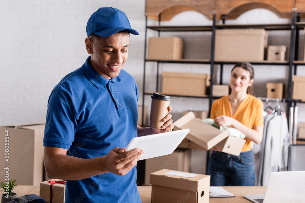 Positive african american deliveryman holding coffee to go and digital tablet near blurred proprietor in online web store. Positive african american deliveryman holding coffee to go and digital tablet near blurred proprietor in online web store.