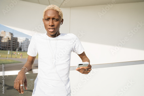 Portrait of smiling confident African American man holding mobile phone looking at camera on unban street. Hipster guy with stylish hair wearing white casual t shirt listening music, copy space  photo