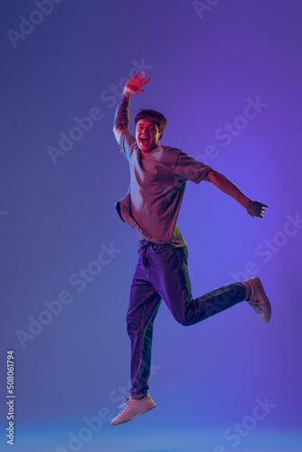 Young red-headed man, student jumping isolated over blue studio background in neon light. Concept of youth, fashion, emotions, facial expression
