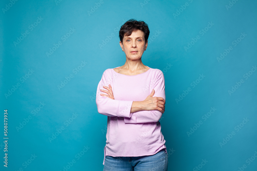 Confident serious mature woman with crossed arms standing against blue studio wall background