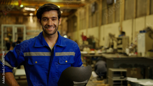 Portrait of smiling male welder in a uniform holding a metal protective mask while working at industrial plant or factory