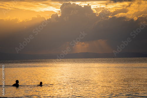 silhouette shot of people swimming under the golden sunlight god rays with clouds surrounding the sun and the blue gold water with waves lapping the shore showing peaceful life at havelock andaman isl photo