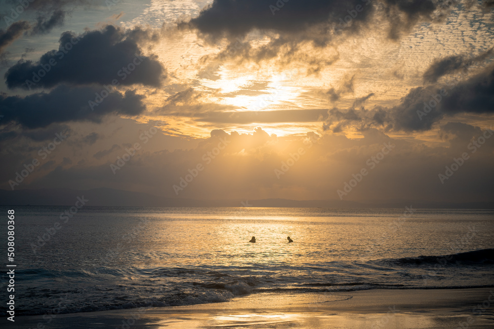 silhouette shot of people swimming under the golden sunlight god rays with clouds surrounding the sun and the blue gold water with waves lapping the shore showing peaceful life at havelock andaman isl