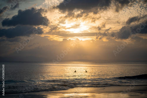 Fototapeta Naklejka Na Ścianę i Meble -  silhouette shot of people swimming under the golden sunlight god rays with clouds surrounding the sun and the blue gold water with waves lapping the shore showing peaceful life at havelock andaman isl