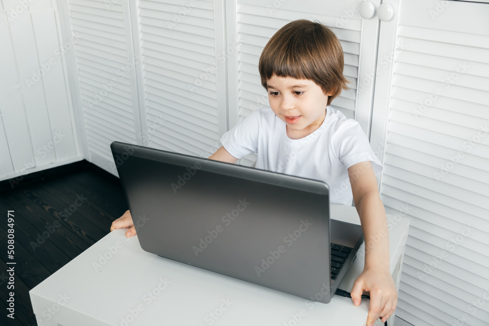 Boy sits at the table, uses laptop, typing on a keyboard, looks at the screen. Child does homework lesson, plays video game, study IT course. Home distance online education concept. Smart technology Boy sits at the table, uses laptop, typing on a keyboard, looks at the screen. Child does homework lesson, plays video game, study IT course. Home distance online education concept. Smart technology