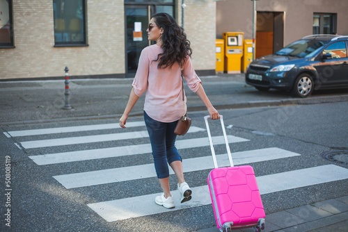 Beautiful young curly woman with a pink small suitcase is in a hurry to go on vacation. A model with long hair crosses the road in a European city. Travels.