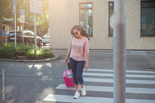Beautiful young curly woman with a pink small suitcase is in a hurry to go on vacation. A model with long hair crosses the road in a European city. Travels.