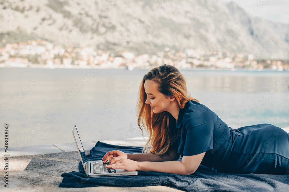 Girl freelancer lies on the background of the mountains with a laptop, remote work. A woman looks at the screen and writes something on the beach in the summer