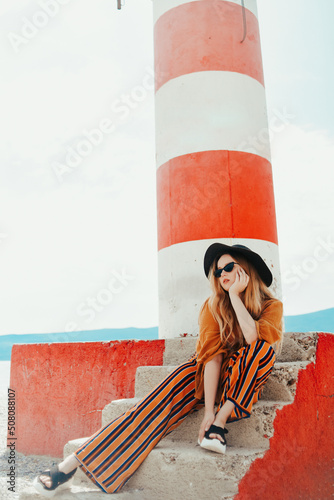 Gorgeous fashionable woman posing while sitting on a lighthouse. Summer photo of a girl in a yellow-orange suit, black hat and sunglasses. Blonde girl posing in travel. photo