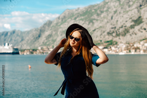 A woman in sunglasses and a hat stands against the backdrop of the sea and mountains, smiling. Vacation and outdoor recreation. Girl with long hair on vacation. Fashion.