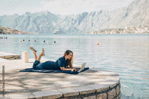 Girl freelancer lies on the background of the mountains with a laptop, remote work. A woman looks at the screen and writes something on the beach in the summer