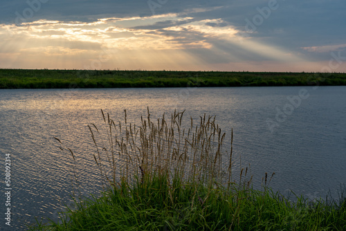 Fototapeta Naklejka Na Ścianę i Meble -  Sunset over the lake. There are small ripples on the water. Reeds in the foreground. The rays of the sun break through the clouds.