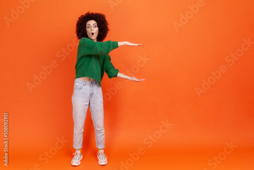 Foto Full length portrait of woman with Afro hairstyle wearing green casual style sweater showing huge size of imaginary subject, copy space