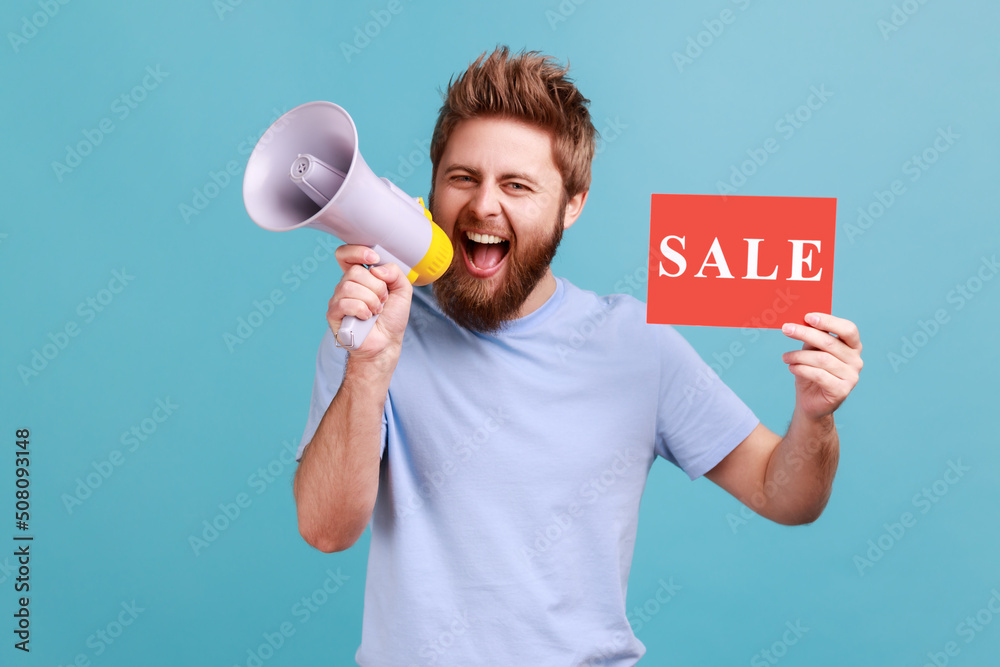 Portrait of positive optimistic handsome bearded man holding card with sale inscription and screaming in megaphone about discounts. Indoor studio shot isolated on blue background. Portrait of positive optimistic handsome bearded man holding card with sale inscription and screaming in megaphone about discounts. Indoor studio shot isolated on blue background.