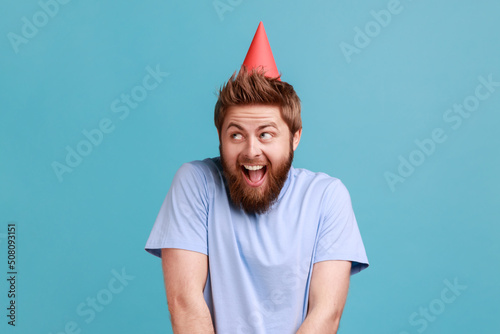 Print op canvas Portrait of pleasant surprised handsome bearded man standing happy facial expression, celebrating her birthday with party cone on his head