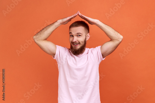 I'm in safety. Portrait of bearded man raising hands showing roof gesture and smiling contentedly, dreaming of house, wearing pink T-shirt. Indoor studio shot isolated on orange background.