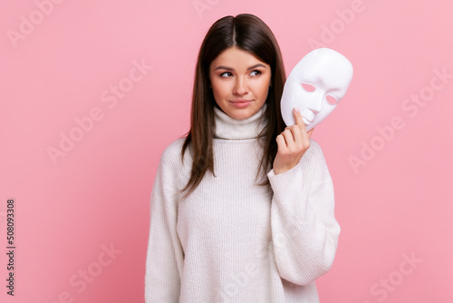 Portrait of young beautiful adult female with dark hair looking away, holding in hands white mask, wearing white casual style sweater. Indoor studio shot isolated on pink background. photo