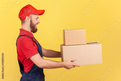 Side view portrait of bearded courier handing cardboard parcels to client, carrying boxes with goods ordered online, wearing blue overalls and red cap. Indoor studio shot isolated on yellow background