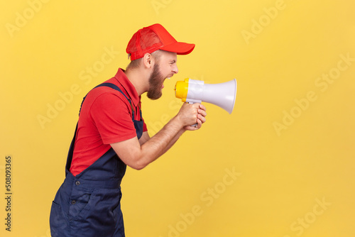 Side view portrait of angry bearded worker in blue uniform and red cap, holding megaphone and screaming with aggressive expression, protesting. Indoor studio shot isolated on yellow background.
