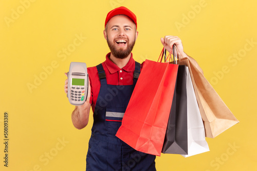 Cheerful joyful delivery man in uniform holding shopping bags and pos payment terminal in his hands, fast delivery from store, service industry. Indoor studio shot isolated on yellow background.