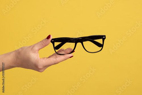 Profile side view closeup of woman hand holding and showing black eyeglasses frame. Indoor studio shot isolated on yellow background.
