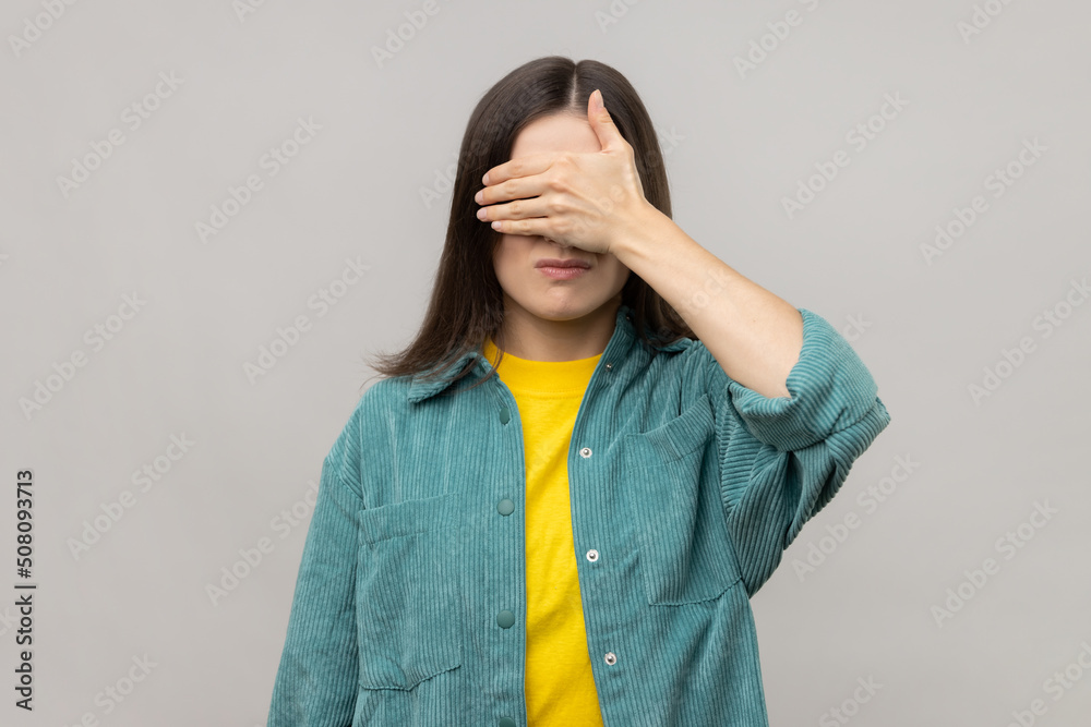 Don't want to look at this. Portrait of shy confused woman covering eyes with hand, feeling shamed and scared to watch, wearing casual style jacket. Indoor studio shot isolated on gray background. Don't want to look at this. Portrait of shy confused woman covering eyes with hand, feeling shamed and scared to watch, wearing casual style jacket. Indoor studio shot isolated on gray background.