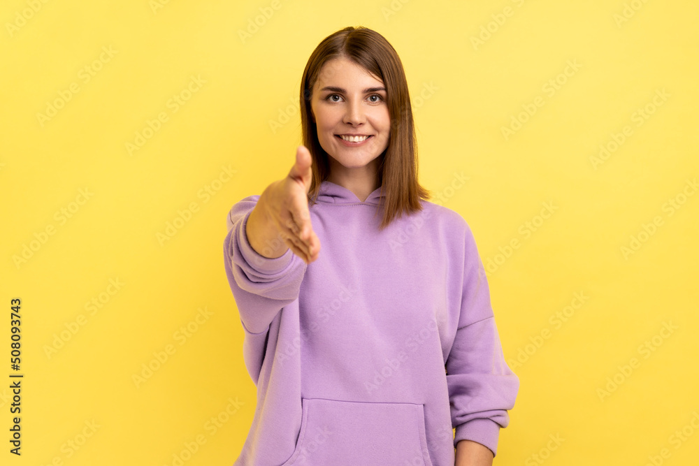 Nice to meet you. Portrait of pleased friendly smiling woman stretching her hand welcoming guests, hospitality, wearing purple hoodie. Indoor studio shot isolated on yellow background. Nice to meet you. Portrait of pleased friendly smiling woman stretching her hand welcoming guests, hospitality, wearing purple hoodie. Indoor studio shot isolated on yellow background.