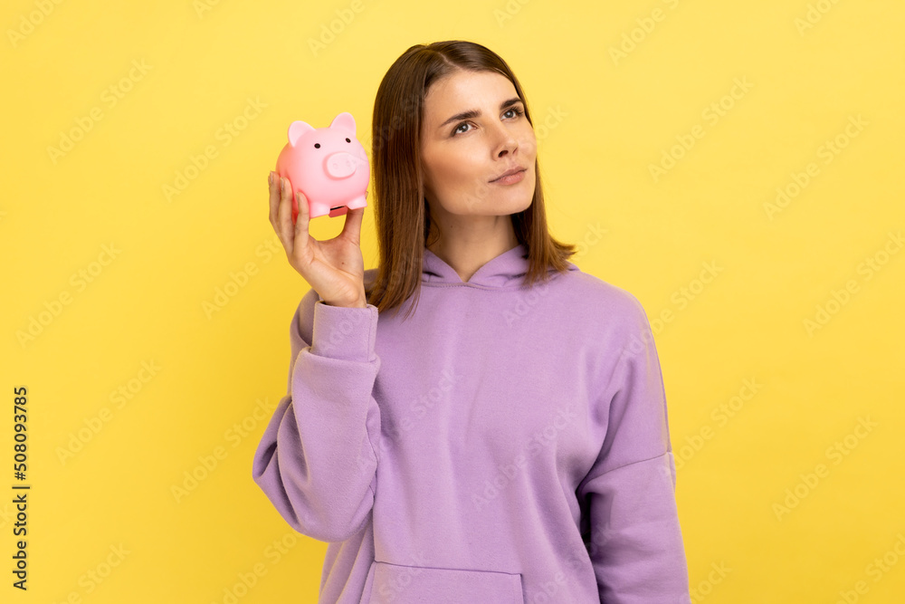 Portrait of pensive woman with pleasant appearance shaking piggy bank, curious of amount of her money savings, wearing purple hoodie. Indoor studio shot isolated on yellow background. Portrait of pensive woman with pleasant appearance shaking piggy bank, curious of amount of her money savings, wearing purple hoodie. Indoor studio shot isolated on yellow background.