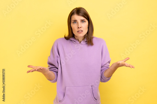 Portrait of dark haired woman raising hands and looking with confused expression, quarreling, annoyed by conflict, wearing purple hoodie. Indoor studio shot isolated on yellow background.