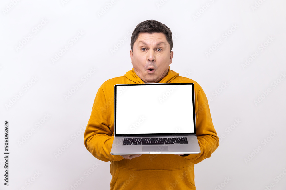 Surprised astonished man holding laptop and looking at camera with open mouth, shocked by new operating system, wearing urban style hoodie. Indoor studio shot isolated on white background.