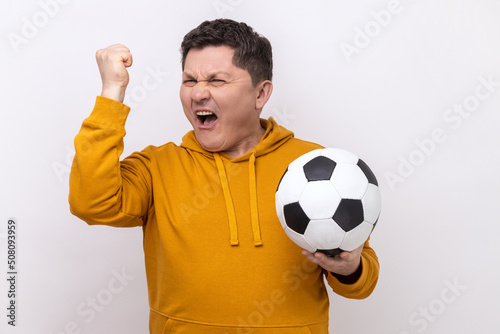 Positive satisfied man screaming holding soccer ball, celebrating victory of favourite football team on championship, wearing urban style hoodie. Indoor studio shot isolated on white background. © khosrork