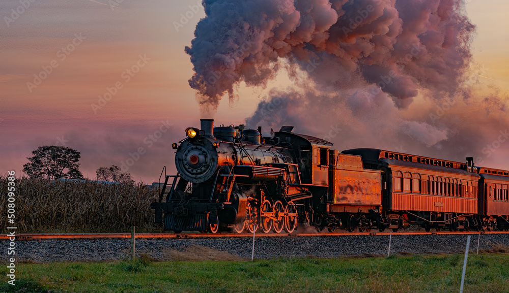 A View of an Antique Steam Passenger Train Approaching at Sunrise With a Full Head of Steam and Smoke Traveling Thru Farmlands