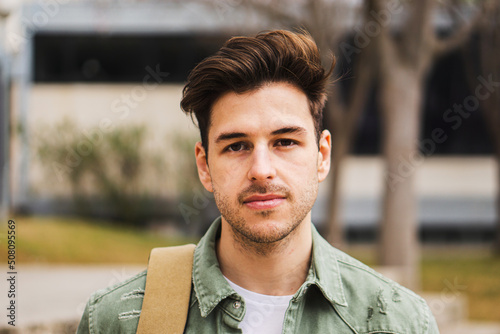 Portrait of teenage male student looking serious at camera in a college. Caucasian guy standing in university campus.
