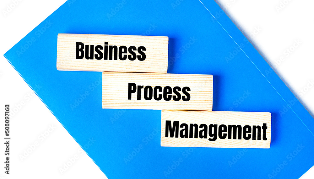 There is a blue notebook on a light gray background. Above are three wooden blocks with the words BPM Business Process Management There is a blue notebook on a light gray background. Above are three wooden blocks with the words BPM Business Process Management
