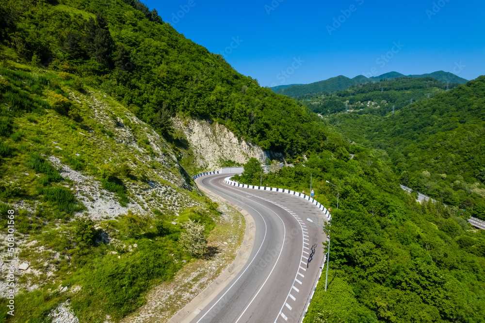 Asphalt road is meandering between blue sea and green mountains. Aerial view of car driving along the winding mountain road in Sochi, Russia.