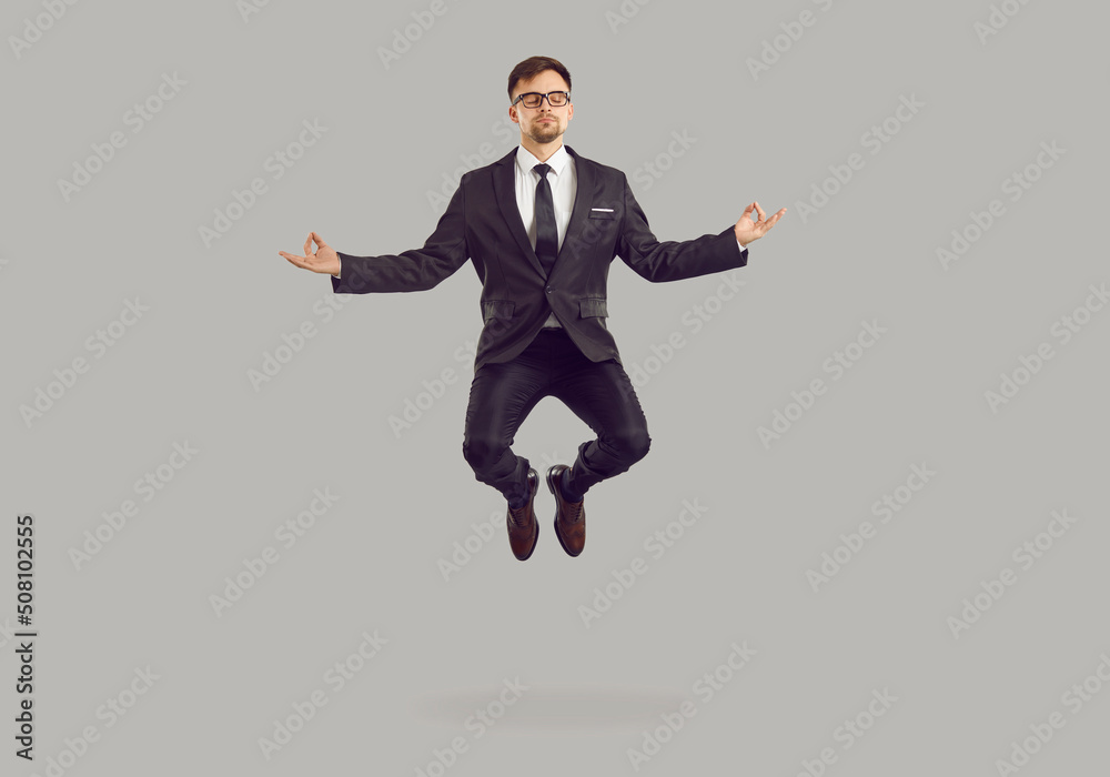 Tranquil male entrepreneur levitating while meditating. Studio portrait of handsome young business man in suit and glasses practising meditation exercise, reaching zen and floating in air in yoga pose