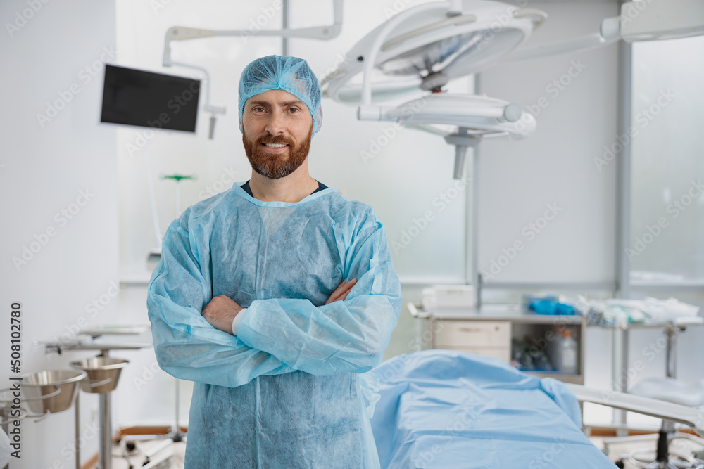 Smiling male surgeon standing in operating room with crossing hands, ready to work on patient Smiling male surgeon standing in operating room with crossing hands, ready to work on patient