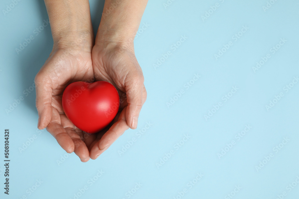 Fototapeta premium Elderly woman holding red heart in hands on light blue background, top view. Space for text
