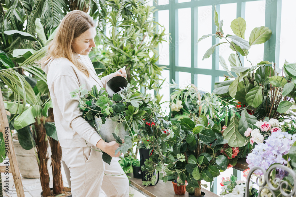 young woman waters flowers from watering can in greenhouse with tropical plants and potted plants, spring concept of urban and home jungles, place for rest and relaxation mental health