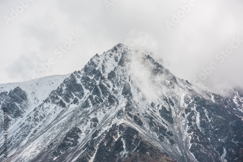 Awesome landscape with high snowy mountain peaked top with sharp rocks in low clouds. Dramatic view to snow mountain pointed peak in low cloudy sky. Monochrome scenery with white snow on black rocks. © Daniil
