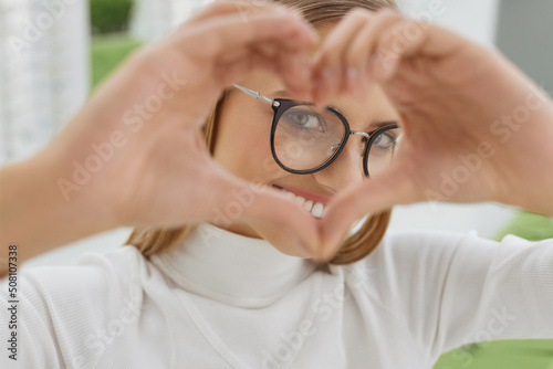 Women Fashion Glasses. Happy Female In Black Optical Frame, Eyewear. Portrait Of Beautiful Young Woman in Stylish Eyeglasses Showing Heart Shape with Hands at Optical Store
