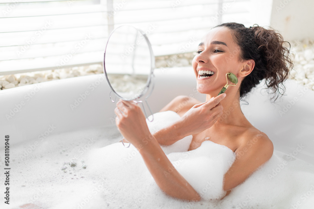 Pretty young woman making facial massage with jade roller near mirror while lying in foamy bath at hotel, copy space Pretty young woman making facial massage with jade roller near mirror while lying in foamy bath at hotel, copy space