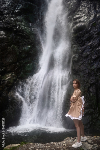 Attractive girl in a beige dress is posing on the background of the waterfall.