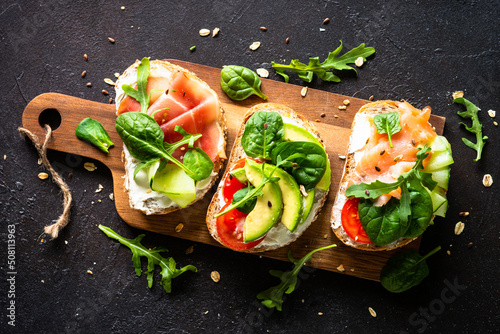 Open sandwich set with cream cheese, prosciutto, salmon, avocado and fresh greens. Top view at dark.