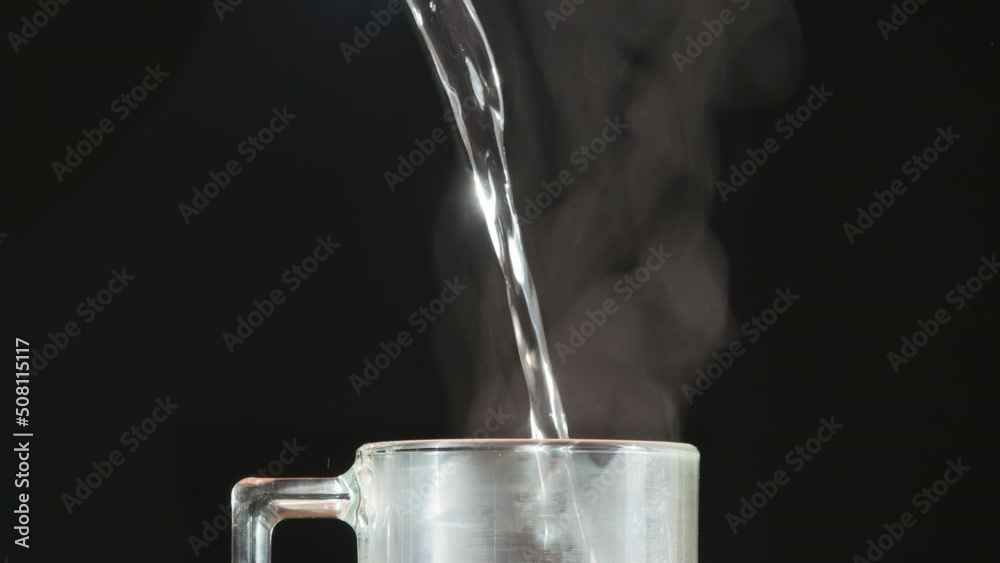 Boiling Water Jet for Tea. A jet of boiling water falls into a glass tea mug, creating puffs of white steam against a black background