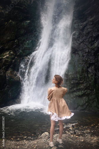 Fresh natural picture of a young beautiful girl in a dress on the background of a waterfall in summer as wallpaper  magazine cover  poster or advertising design. Summer happiness vibe.