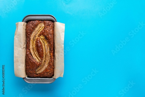 Traditional American homemade banana bread with chopped walnuts, chocolate chips and cinnamon in loaf pan on blue background. Fruit cake. Healthy vegan desserts concept. Top view, copy space.
