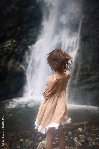 Slim brunette girl in a beige dress is on the background of the waterfall.